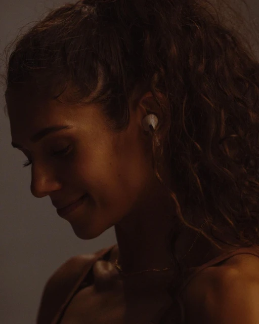 Side view of a smiling adult woman with curly hair and wireless earbuds, softly lit against a neutral studio backdrop.