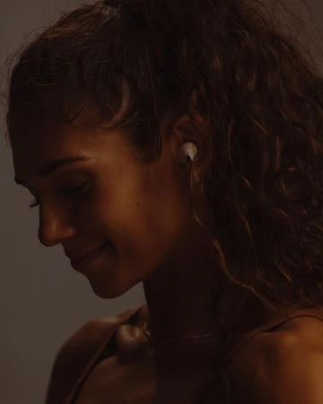 Side view of a smiling adult woman with curly hair and wireless earbuds, softly lit against a neutral studio backdrop.