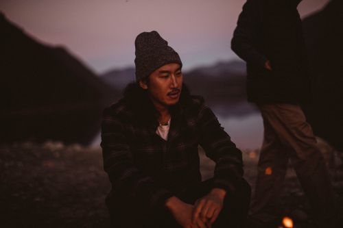 Asian man wearing a beanie and jacket sits near a campfire at dusk with another person visible and mountains in the background.