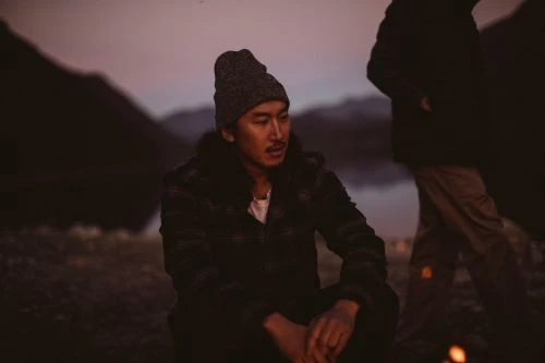 Asian man wearing a beanie and jacket sits near a campfire at dusk with another person visible and mountains in the background.