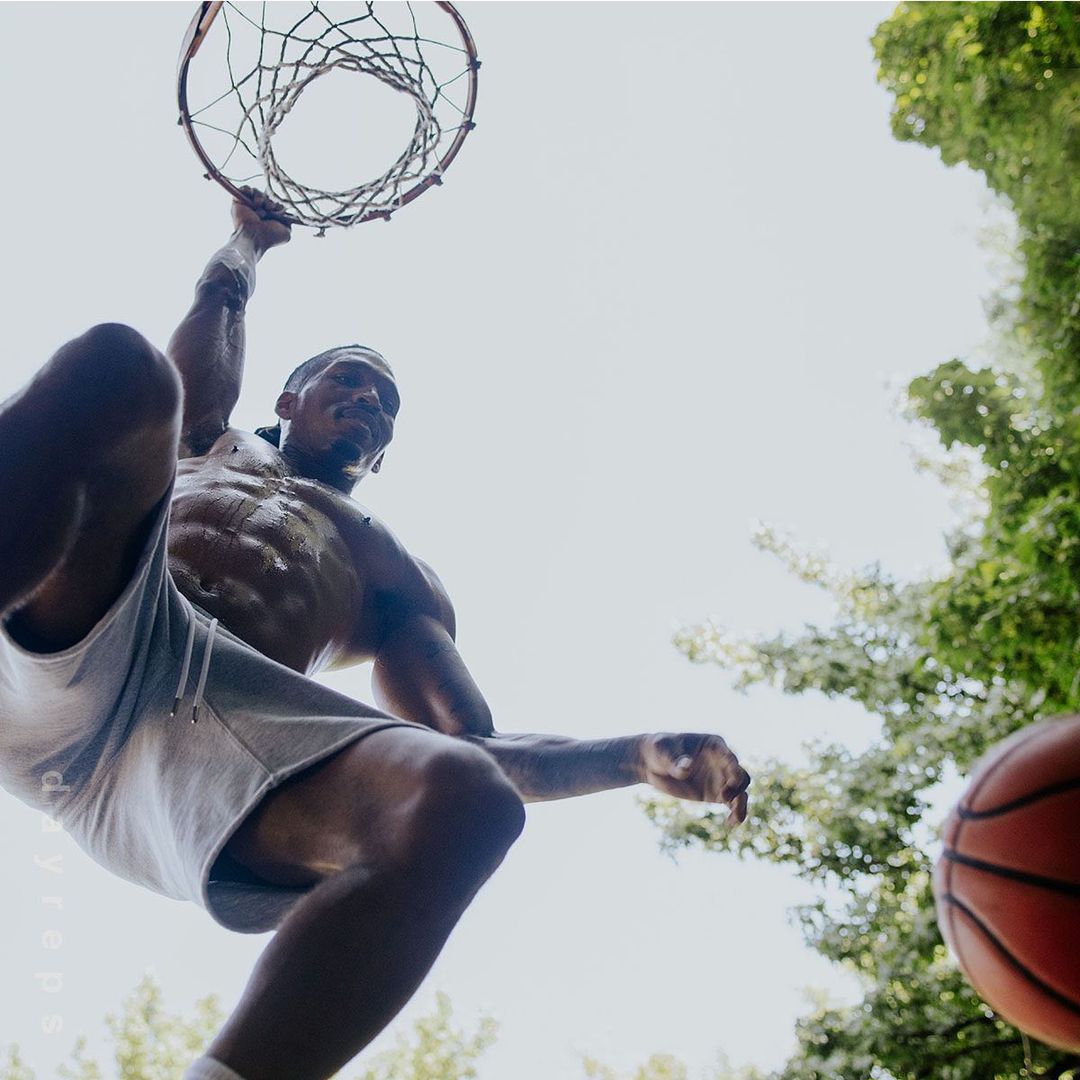 Low angle of muscular man dunking basketball outdoors with green trees in the background. Upward perspective.