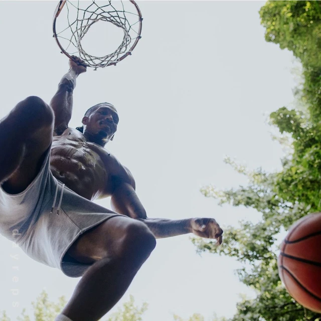 Low angle of muscular man dunking basketball outdoors with green trees in the background. Upward perspective.