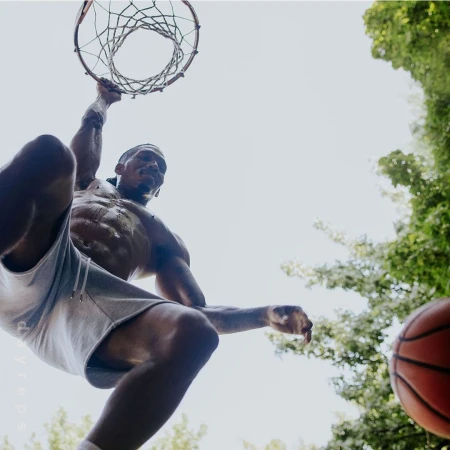 Low angle of muscular man dunking basketball outdoors with green trees in the background. Upward perspective.