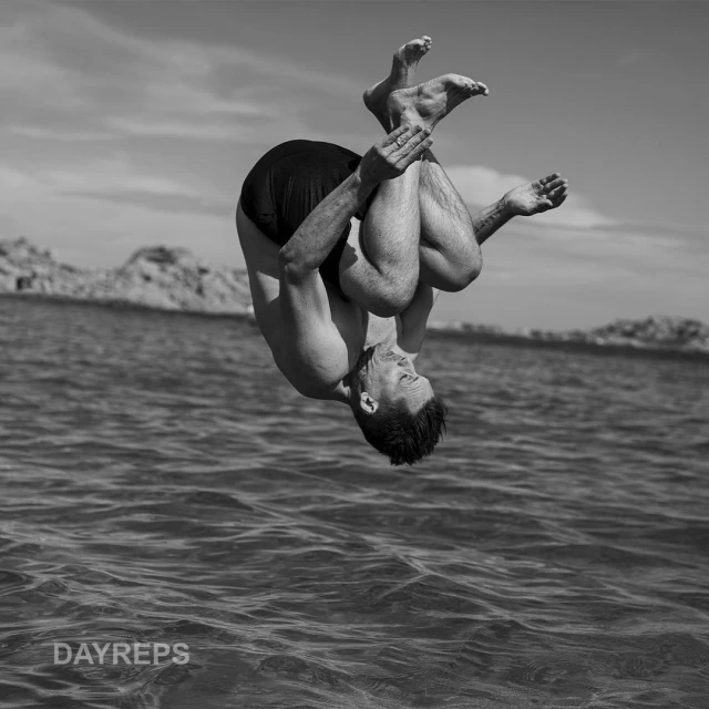 Black and white photo of an adult male summersaulting over the water, with land and sky in the background, and a visible watermark.