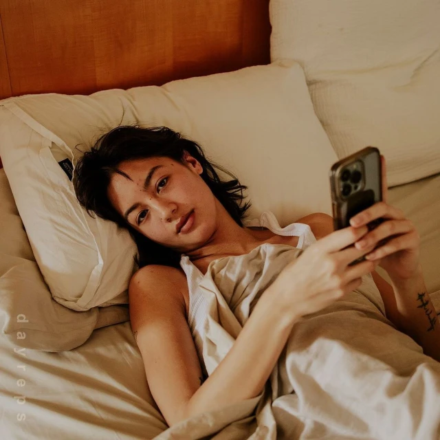 A woman lies in bed holding her cell phone. She is indoors, resting on pillows and under bedsheets in soft, neutral colors.