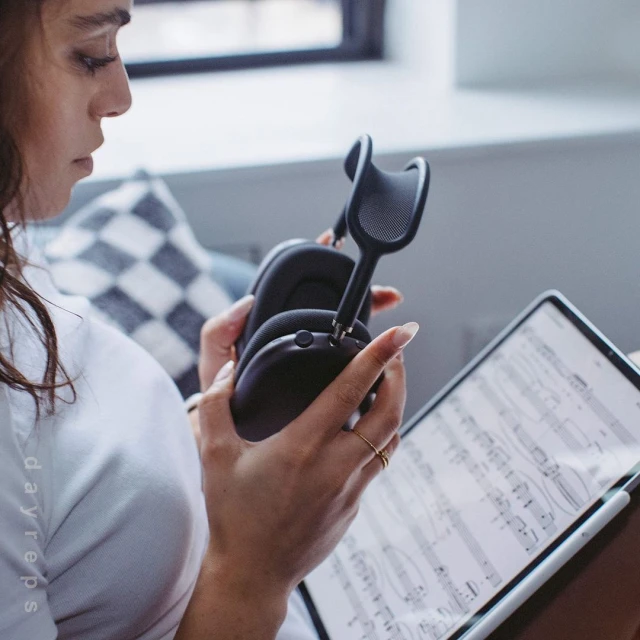 A woman holds black headphones while working on a tablet displaying sheet music in a brightly lit room by a window.
