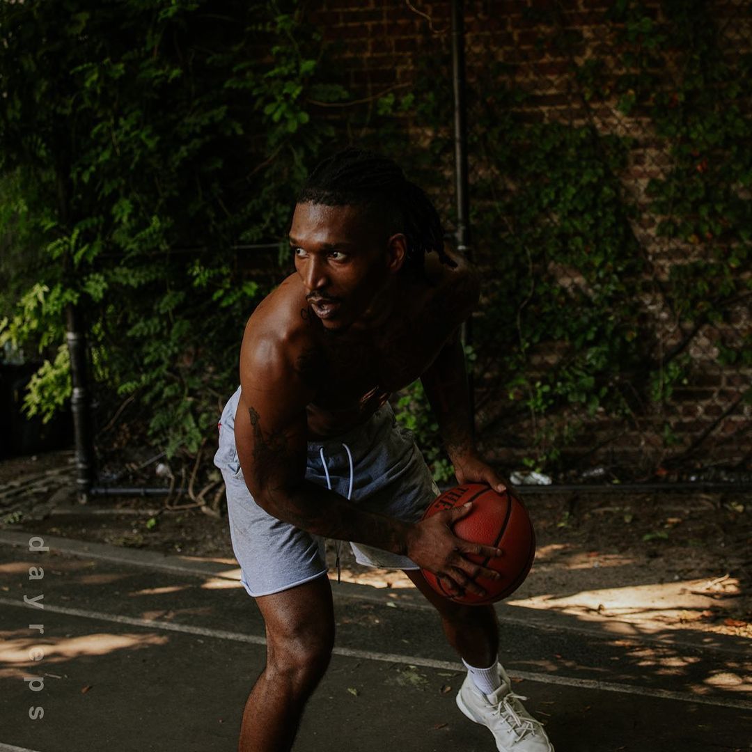 Athletic Black man dribbling a basketball on an outdoor court with a brick wall and green vines in the background on a sunny day.