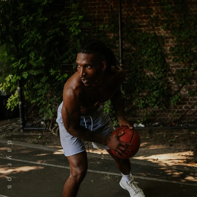 Athletic Black man dribbling a basketball on an outdoor court with a brick wall and green vines in the background on a sunny day.