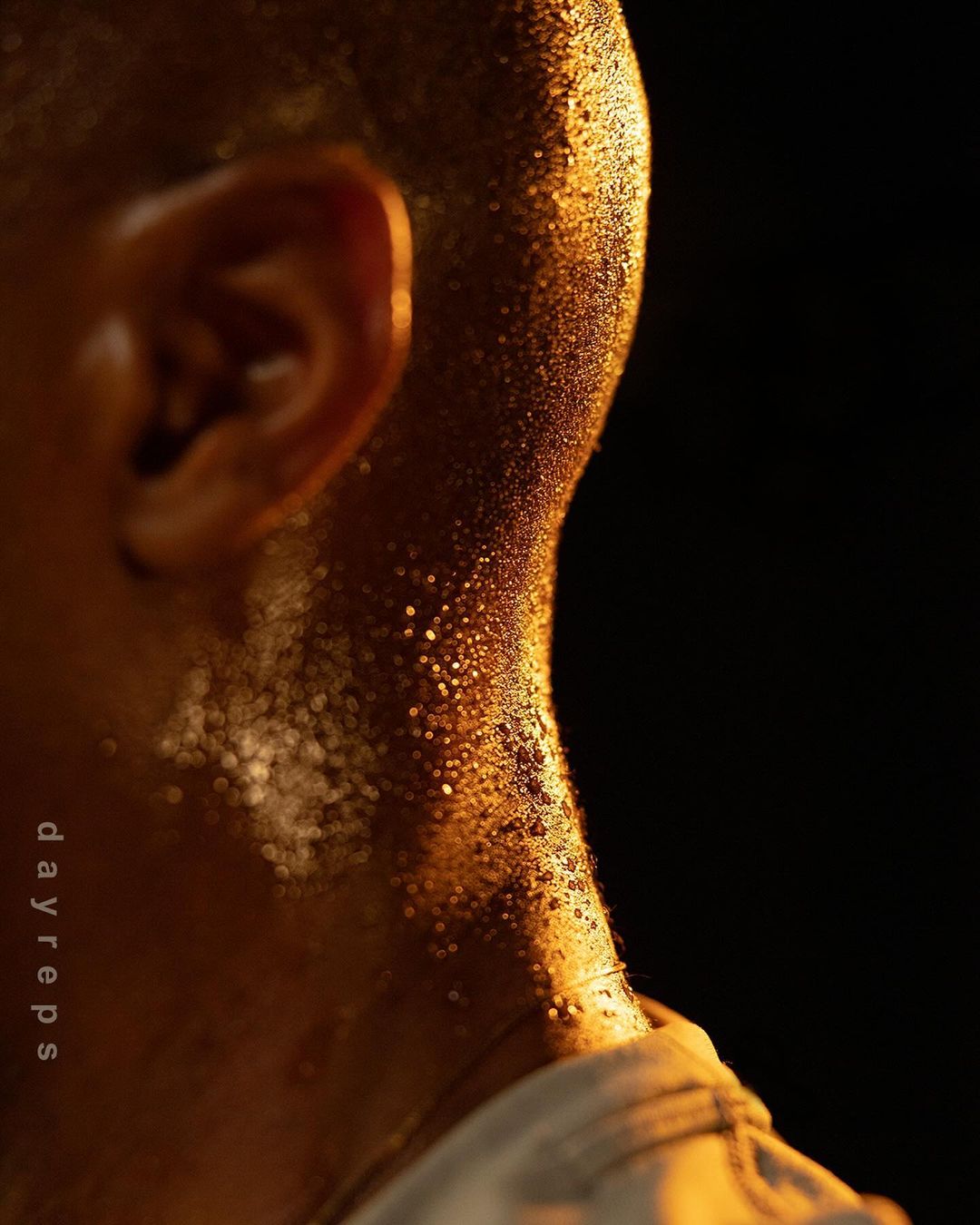 Close-up of a person's neck and ear covered in sweat droplets, reflecting warm light against a dark background, implying a workout or physical activity.