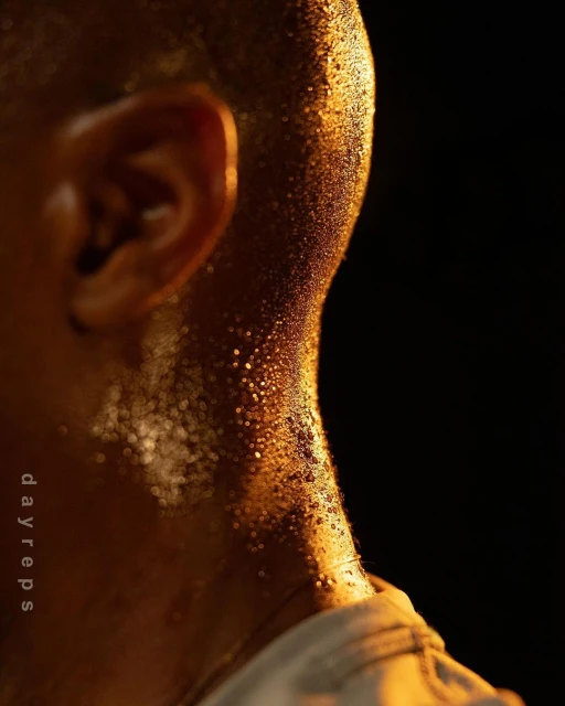 Close-up of a person's neck and ear covered in sweat droplets, reflecting warm light against a dark background, implying a workout or physical activity.