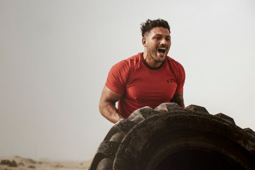 Muscular man in red t-shirt flipping a large tire outdoors in a fitness setting, showcasing strength and determination in sports training.
