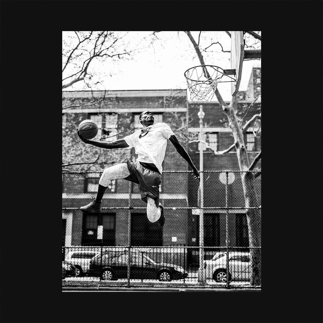 Black and white image of a Black adult male leaping toward a basketball hoop with ball in hand on an outdoor basketball court.