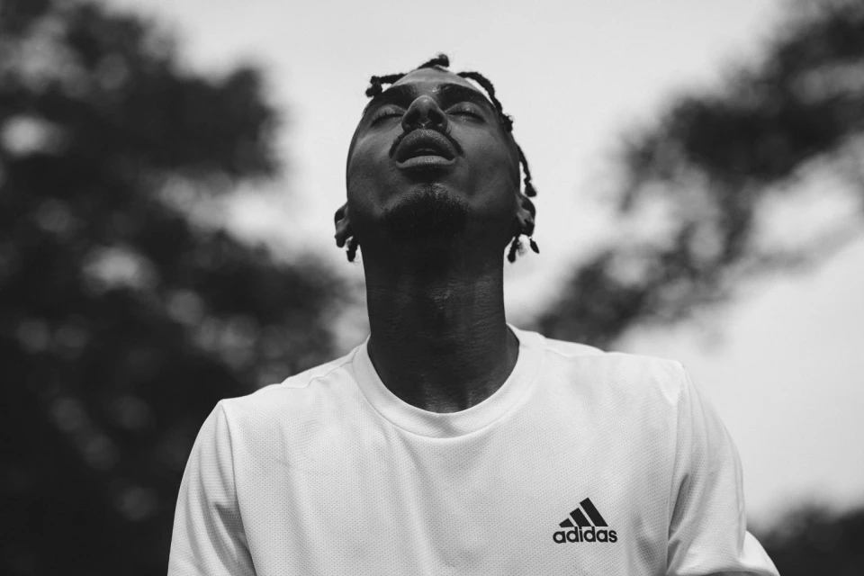Black and white portrait of an adult male with locs, eyes closed, wearing a white Adidas t-shirt against an outdoor, blurry background.