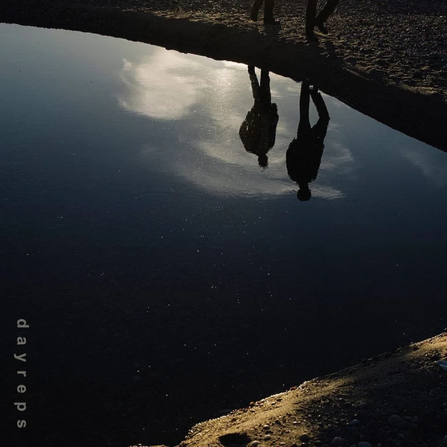 Low-angle view of two silhouetted figures reflected in the dark water, surrounded by sandy land, creating a mirrored effect.