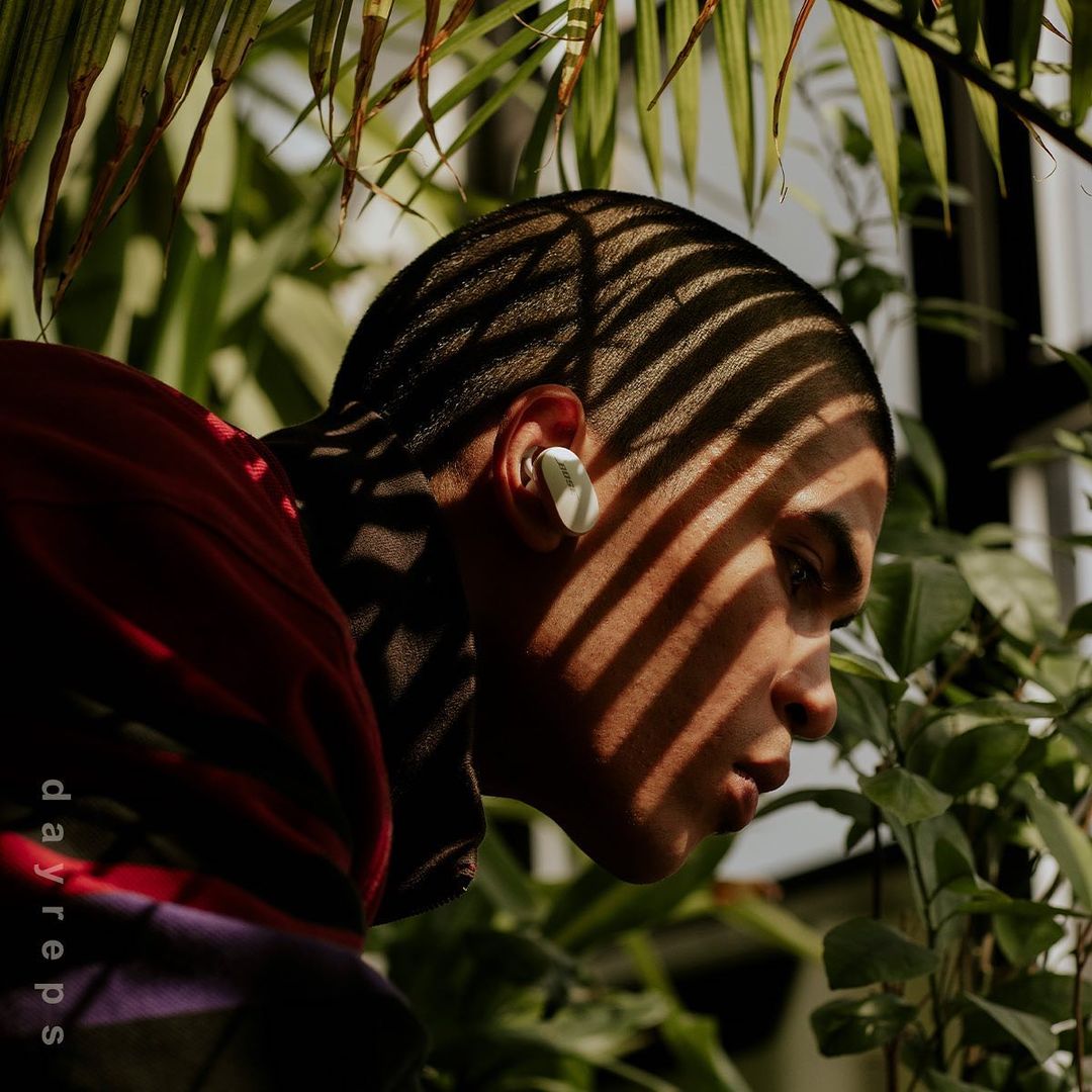 Close-up of a person with earbud in ear surrounded by green foliage. Light and shadow play across the subject's face.