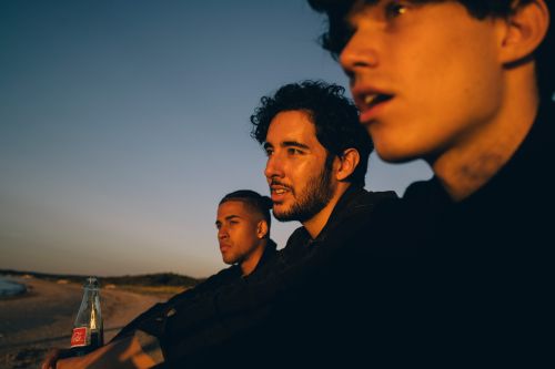Three young adult men sit near a beach at dusk, silhouetted against a twilight sky, with a glass soda bottle nearby, creating a casual atmosphere.