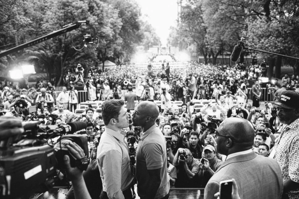 Two men face each other closely in a public square surrounded by a large crowd with cameras, booms, and recording equipment.