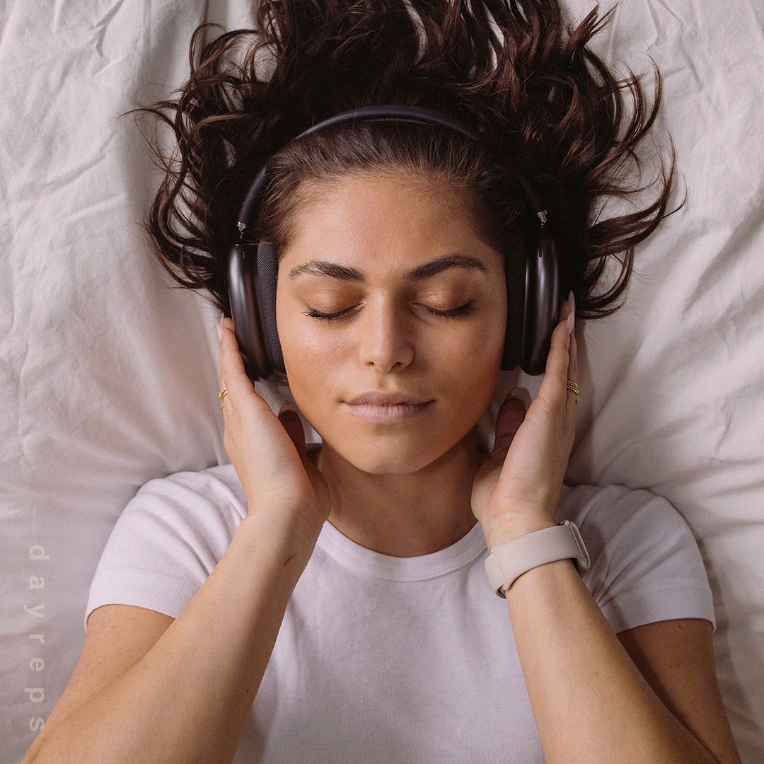 Woman laying on bed with headphones. Young adult female with dark hair relaxing, wearing headphones, eyes closed on a white bed sheet.