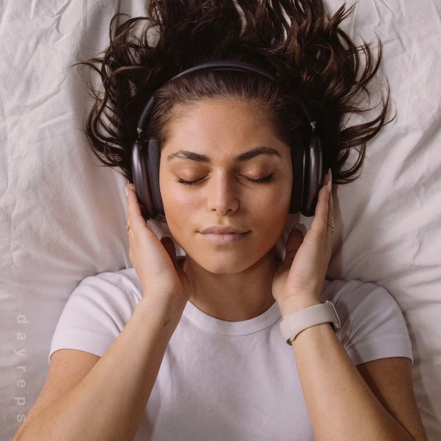 Woman laying on bed with headphones. Young adult female with dark hair relaxing, wearing headphones, eyes closed on a white bed sheet.
