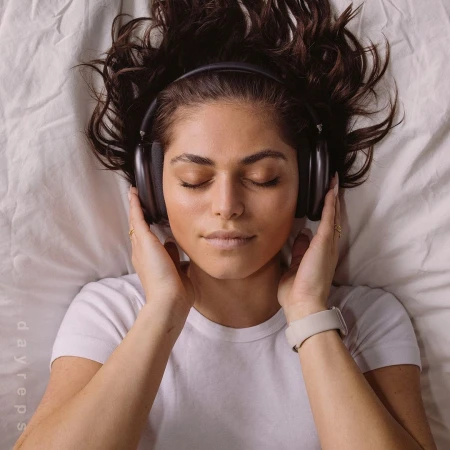 Woman laying on bed with headphones. Young adult female with dark hair relaxing, wearing headphones, eyes closed on a white bed sheet.