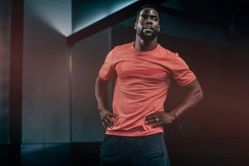 Close-up of a fit African-American adult male in a red t-shirt posing indoors with hands on hips, set against a blurred background.