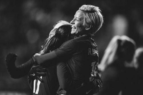 Two female soccer players embrace and smile on a field, captured in black and white, showcasing sportsmanship and celebration.