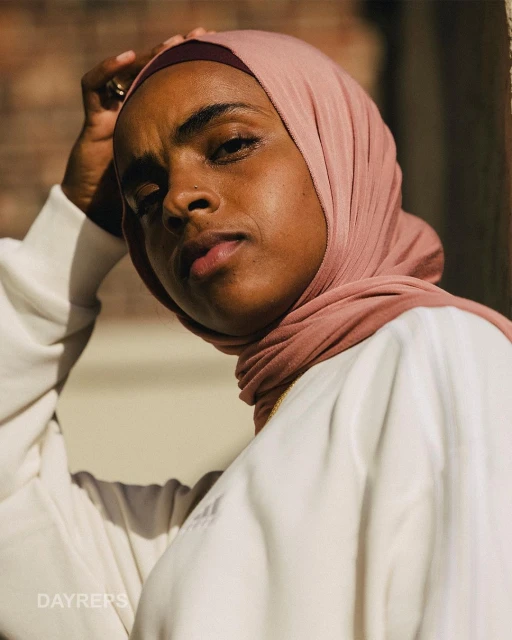 Portrait of a beautiful woman wearing a pink headscarf and white long-sleeve shirt posing outdoors with natural, soft light.