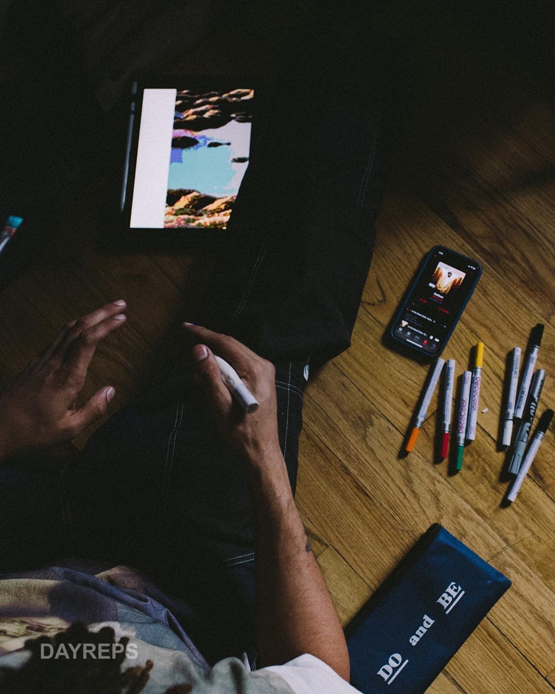 Person using a digital pen with a tablet on a wood floor next to colored markers, a phone, and a case with text. 
