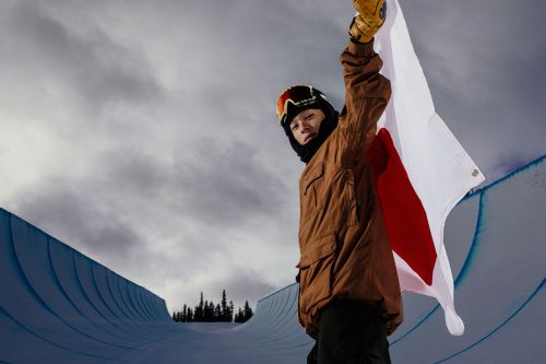 Young snowboarder holds the Japanese flag on a snow halfpipe against a cloudy sky, wearing a brown jacket and goggles.