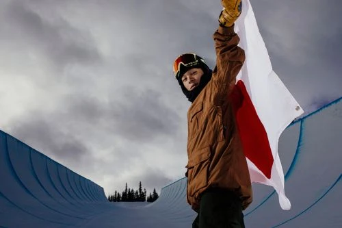 Young snowboarder holds the Japanese flag on a snow halfpipe against a cloudy sky, wearing a brown jacket and goggles.