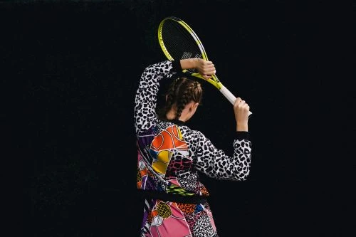 Back view of a woman holding a tennis racket overhead against a dark background wearing a colorful patterned athletic outfit indoors.