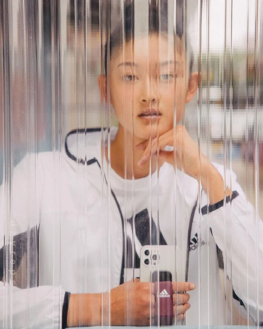 Asian woman with hand to face, posed indoors, wearing white Adidas athletic jacket, holding smartphone, looking through clear plastic curtain.
