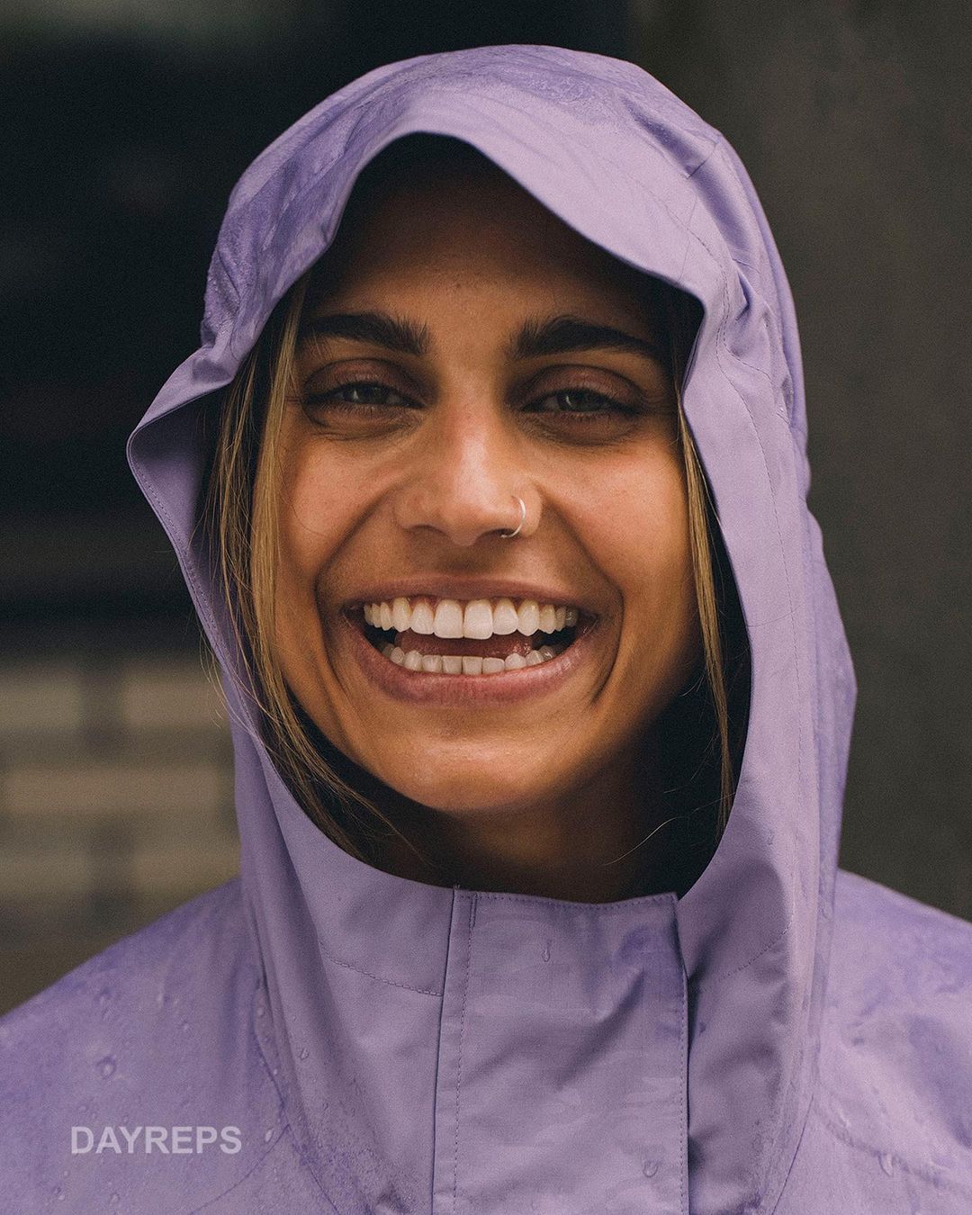Close-up of an adult woman smiling, wearing a lavender hooded jacket. The photograph captures natural light and a happy expression.