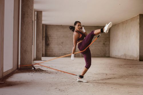 A woman in sportswear wearing ankle weights practices a kick with a resistance band in a concrete building.