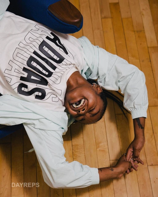 Portrait of a smiling African-American woman in sportswear with the Adidas logo, posing upside down on a wooden surface in casual setting.