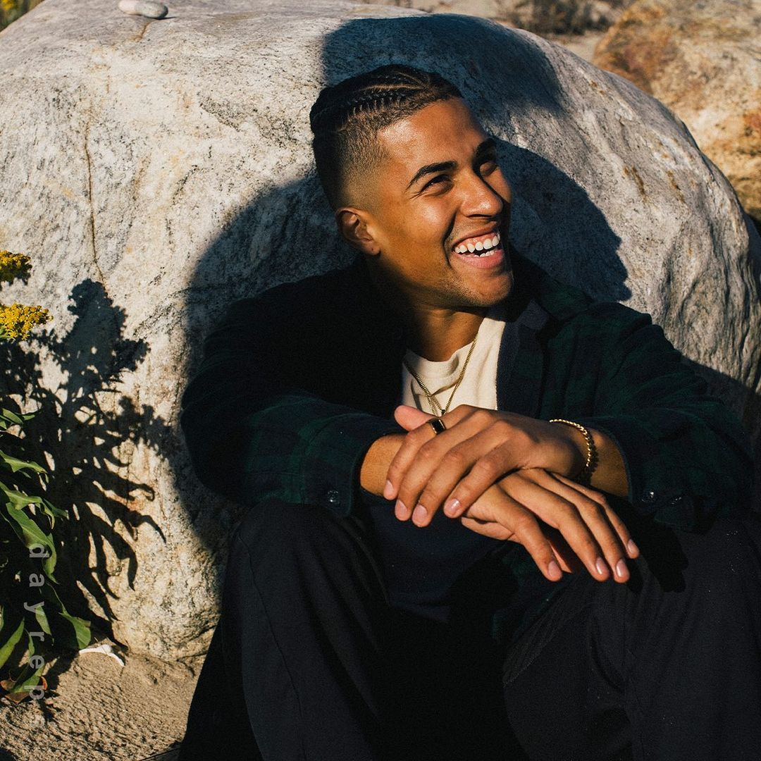 Smiling man with braided hair seated outdoors against a large boulder in Los Angeles, photographed by Nick Laham