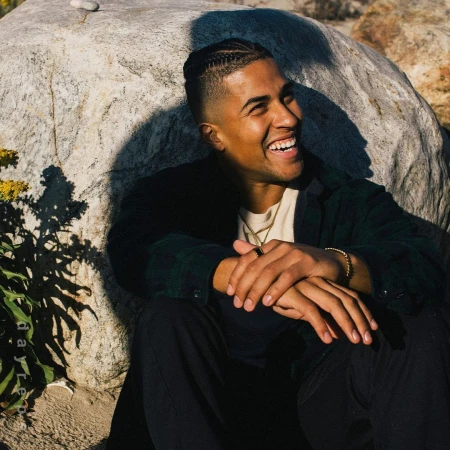 Smiling man with braided hair seated outdoors against a large boulder in Los Angeles, photographed by Nick Laham