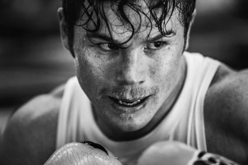 Close-up black and white shot of a sweaty, determined adult male boxer with dark hair and a mustache, wearing a white tank top.