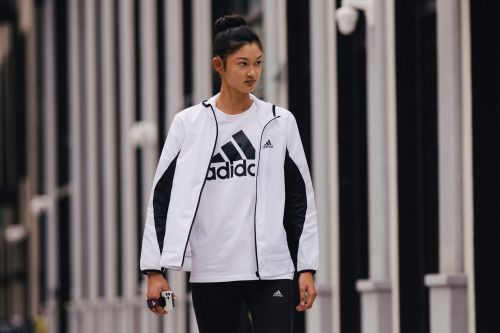 An Asian adult female stands outdoors in a white Adidas jacket and black leggings, holding a phone, against a blurred white and black background.