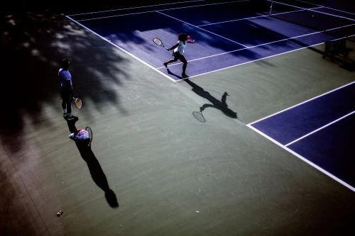 Two tennis players on a blue and green outdoor court in bright sunlight. The player in motion casts a shadow on the court.