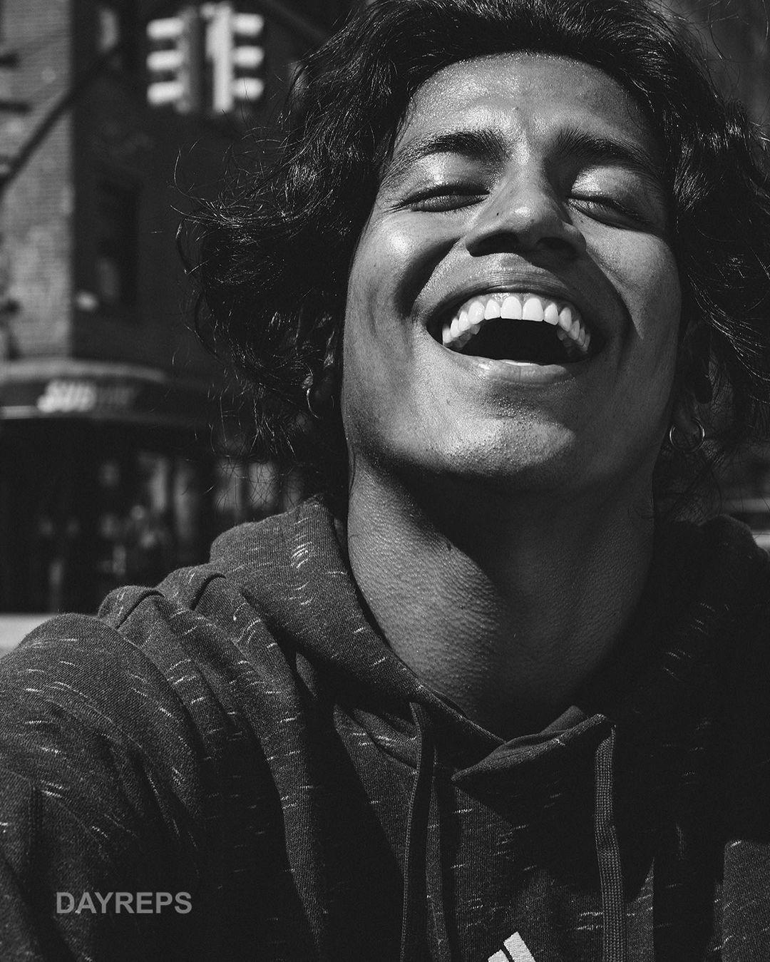 Portrait of a young man with curly hair laughing in an urban setting. He's wearing a hooded sweatshirt, captured in high contrast black and white.