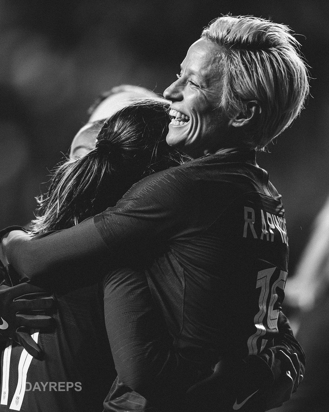 Two smiling athletes in soccer uniforms embrace on a field. The scene is black and white, capturing an emotional moment of celebration.