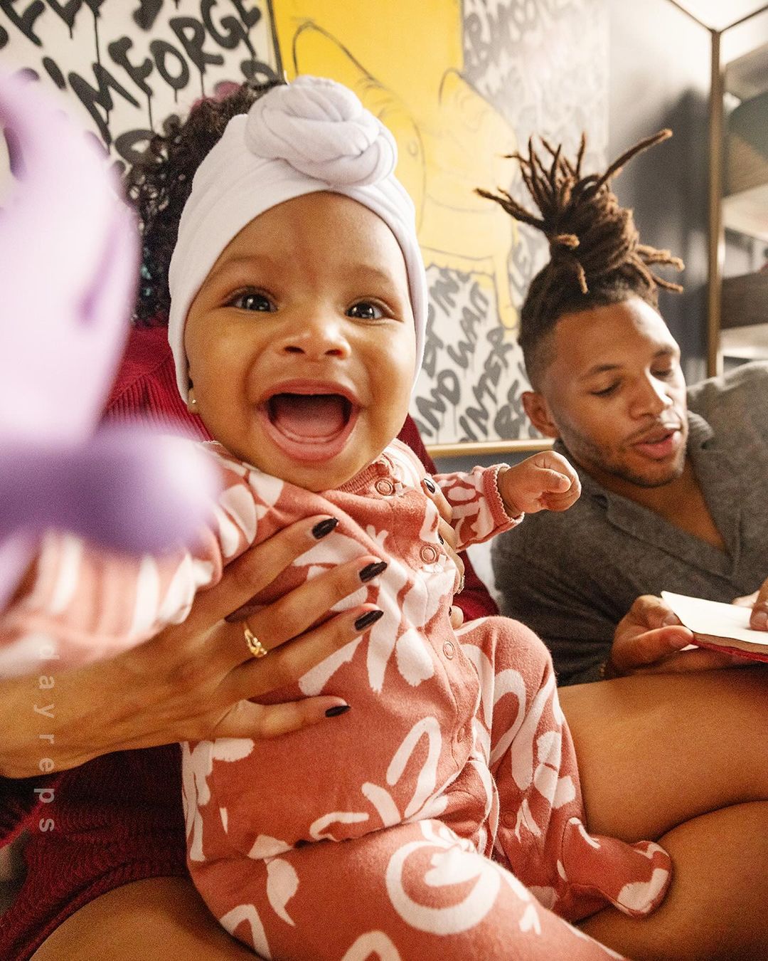 Close-up shot of a smiling baby wearing a head wrap and patterned outfit, held by an adult with an adult male in the background.