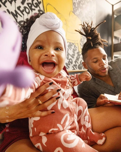 Close-up shot of a smiling baby wearing a head wrap and patterned outfit, held by an adult with an adult male in the background.