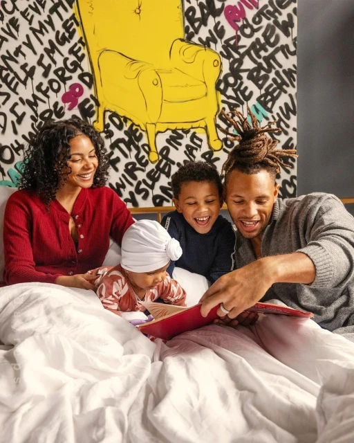 African-American family of four reading a book together in bed in front of a colorful abstract graphic wall. Father, mother, and two children. 