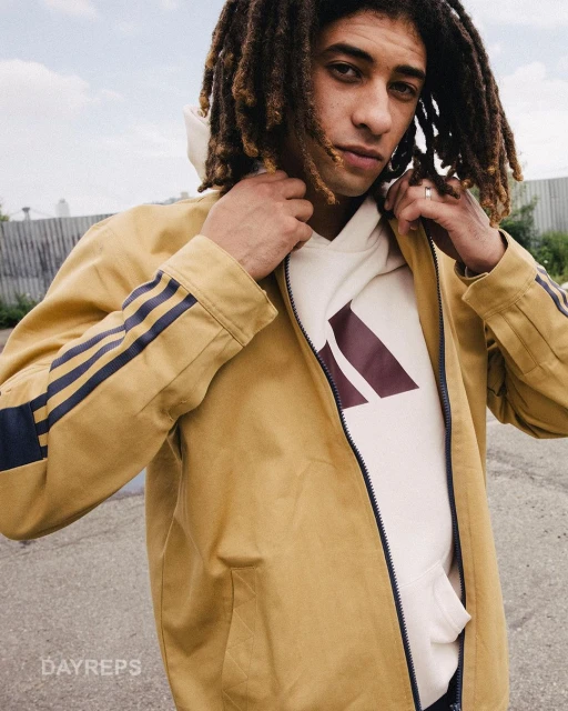 Man with dreadlocks wearing a tan jacket and cream hoodie with logo, outdoors. Casual fashion shot, urban style, medium shot.