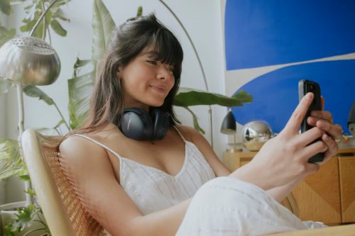 Smiling woman in a white dress sits in a chair indoors, holding a smartphone while wearing black headphones around her neck.