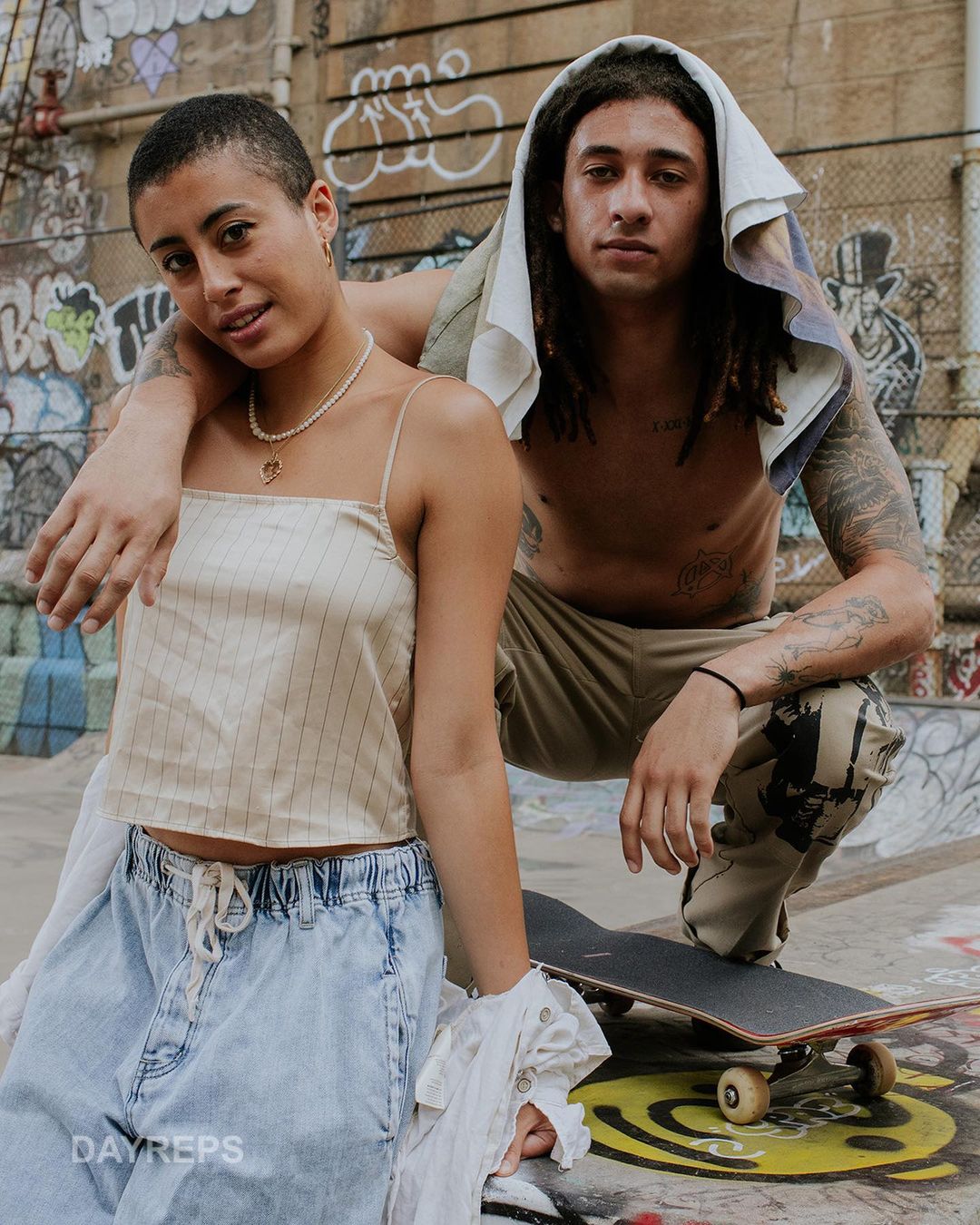 Two young adults pose at a skatepark. A young woman stands next to a crouching young man with tattoos and dreadlocks with a skateboard.