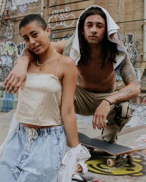 Two young adults pose at a skatepark. A young woman stands next to a crouching young man with tattoos and dreadlocks with a skateboard.