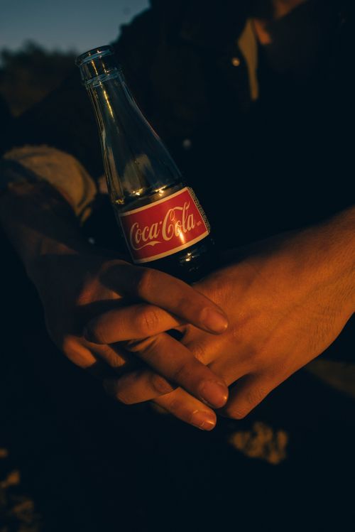 Person holding a glass Coca-Cola bottle. The red Coca-Cola label is visible. The image focuses on the hands, bottle, and dark background.